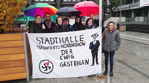 wenige Personen stehen unter Regenschirmen hinter einem Protestbanner gegen die AfD